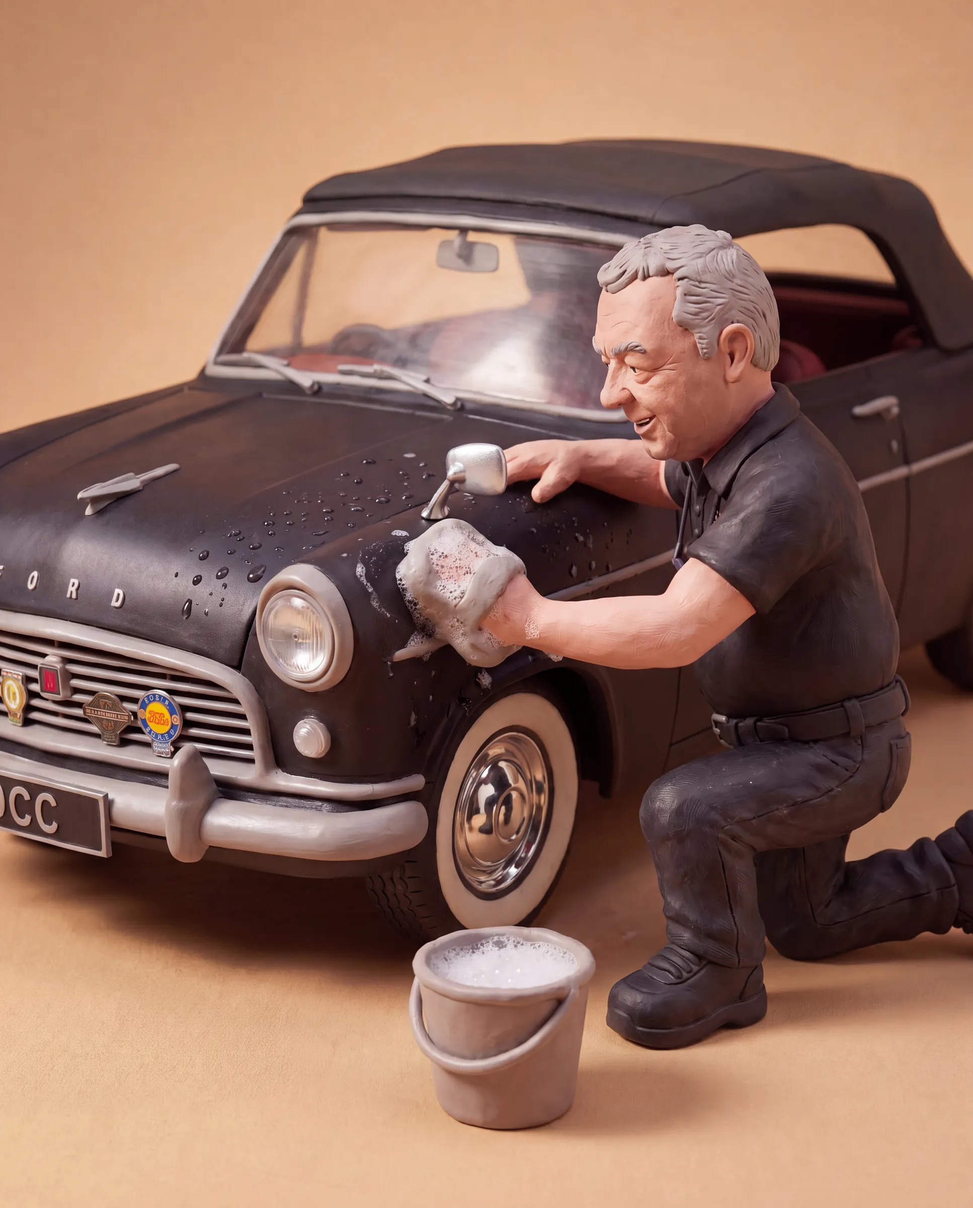 Clay model of Dean kneeling beside his black Ford Consul, hood up, washing the front wing with a soapy sponge, with a bucket of suds on the ground next to him