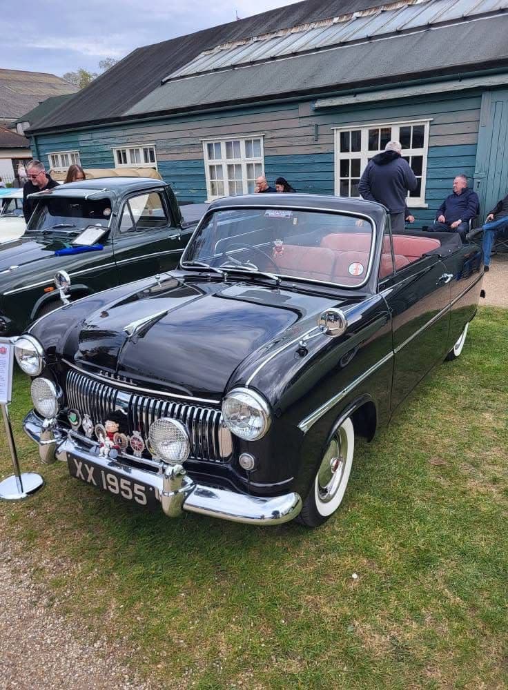 Dean's black Ford Consul drophead with the hood down and red leather seats, parked on the grass beside a weatherboarded building at a Brooklands car show, with people sitting out in the background